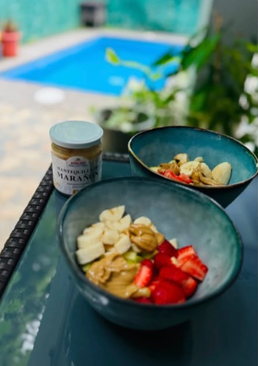Dos tazones con fruta y mantequilla de marañón sobre una mesa, con la piscina detrás